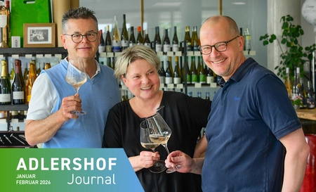 Three people standing in front of a wine rack toast with wine glasses.