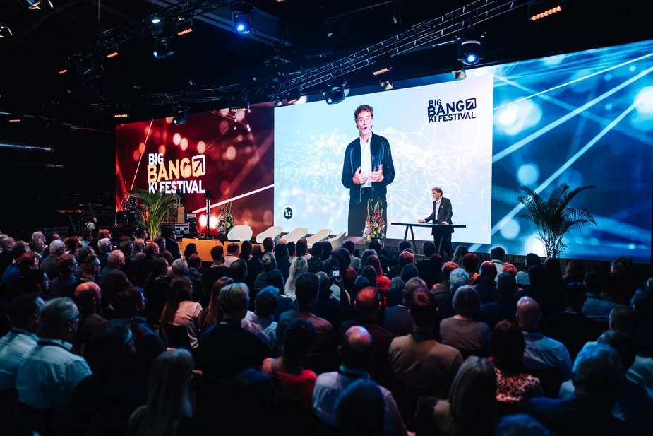 Bühne mit großem Bildschirm und sitzendem Moderator vor Publikum bei einer Veranstaltung mit dem Schriftzug 'BIG BANG KI FESTIVAL'