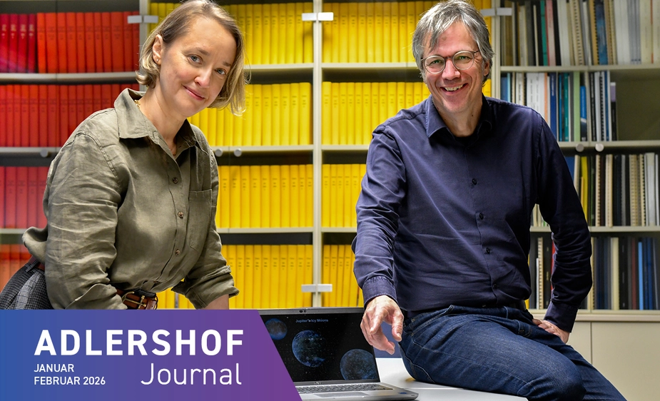 Zwei Menschen stehen an einem Tisch voller Bilder vor einem Bücherregal