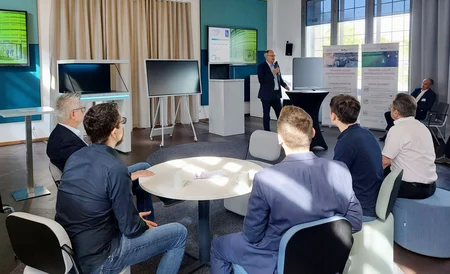 Vertreter der Firmen sitzen um einen runden Tisch oder auf Hockern in der a:head area in Adlershof. Ein Mann im Bildhintergrund präsentiert gerade. Hinter ihm stehen Aufsteller der BTU und Monitore.