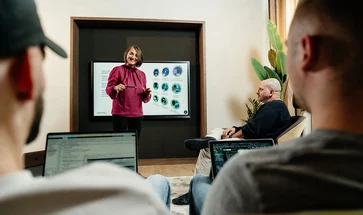 Eine Frau steht vor einem Bildschirm und hält einen Vortrag. Zuhörende sitzen zum Teil mit Laptops auf dem Schoß.