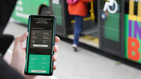 Smartphone in der Hand eines Menschen, im Hintergrund steigt eine Person in einen Bus ein