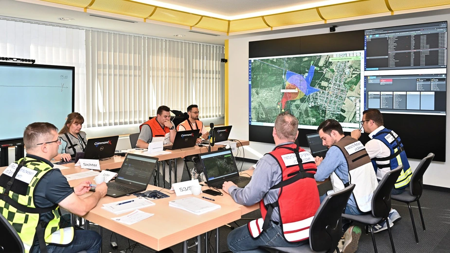 Teilnehmende sitzen in einem Konferenzraum an Laptops, auf dem Monitor ist ein Lageplan der Situation.