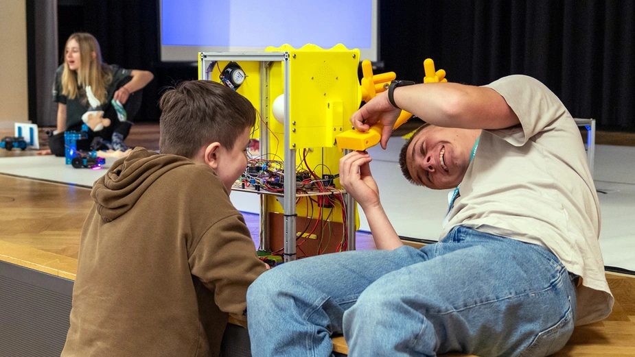 Zwei Schüler schrauben an einem Roboter, der auf der Bühne steht