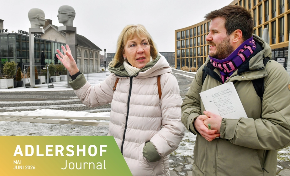 Zwei Personen auf dem winterlichen Platz vor einem modernen Gebäude und einer Skulptur mit zwei Köpfen.