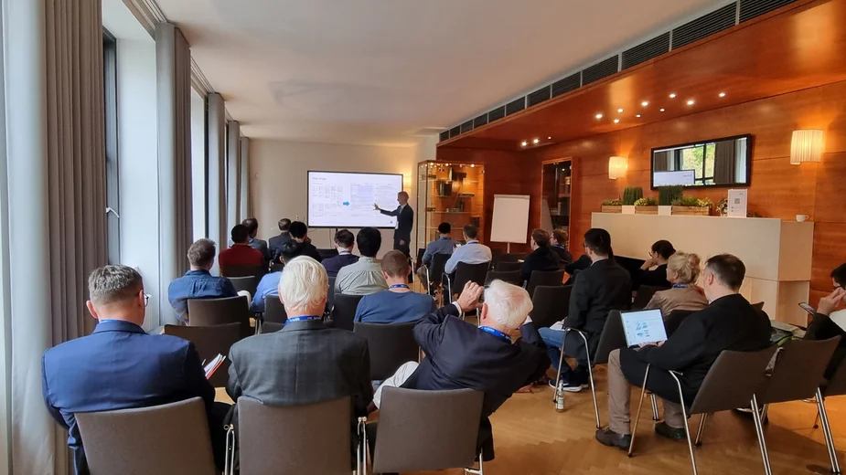 Mehrere Personen sitzen in einem Konferenzraum und hören einem Vortragenden zu, der vor einem Bildschirm steht und auf eine Präsentation zeigt